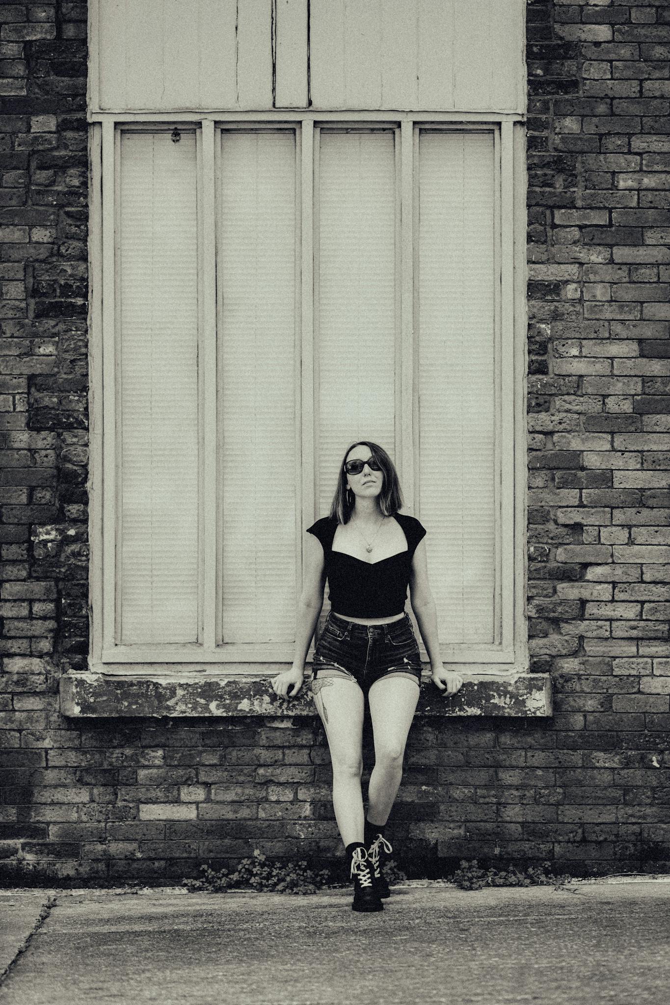 A stylish woman in sunglasses poses against a brick wall with a vintage feel.